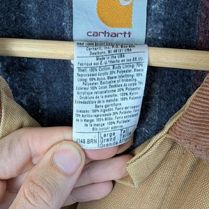 Peut inclure: Gros plan sur l'&eacute;tiquette d'une veste Carhartt. L'&eacute;tiquette est blanche avec du texte noir, comprenant le logo de la marque et la composition du mat&eacute;riau. La veste est marron avec une doublure gris fonc&eacute;.