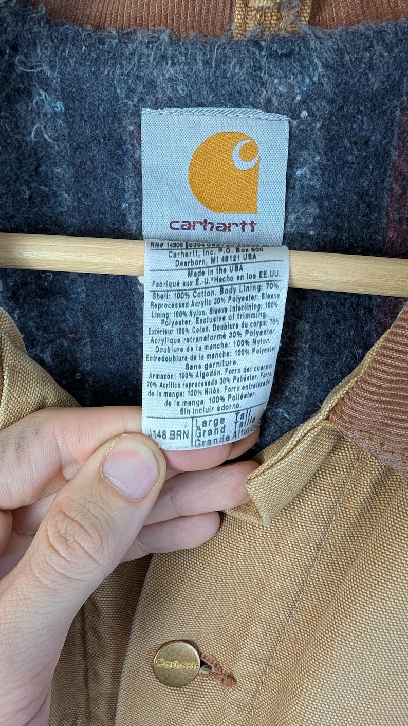 Peut inclure: Gros plan sur l'&eacute;tiquette d'une veste Carhartt. L'&eacute;tiquette est blanche avec du texte noir, comprenant le logo de la marque et la composition du mat&eacute;riau. La veste est marron avec une doublure gris fonc&eacute;.