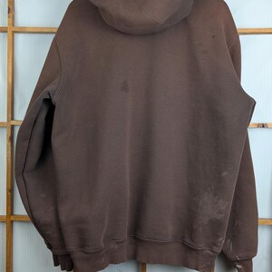 May include: A brown hooded sweatshirt hanging on a wooden hanger. The sweatshirt has a drawstring hood and a kangaroo pocket. There are stains on the front and back of the sweatshirt. The background is a wooden window frame.