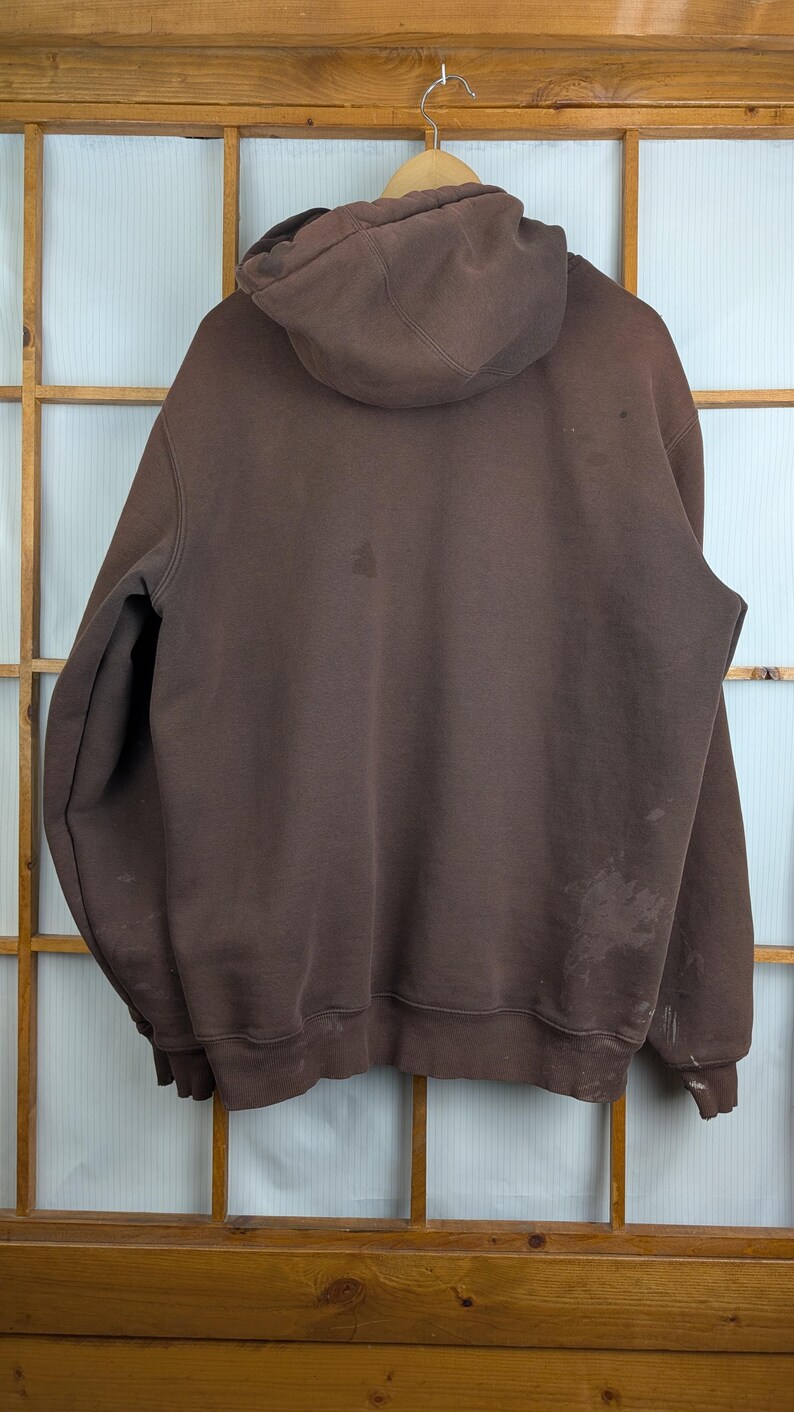 May include: A brown hooded sweatshirt hanging on a wooden hanger. The sweatshirt has a drawstring hood and a kangaroo pocket. There are stains on the front and back of the sweatshirt. The background is a wooden window frame.
