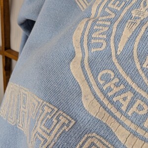 May include: Pale blue sweatshirt featuring a white graphic. The graphic includes the words "UNIVERSITY" and "CHAPEL HILL" in a circular design. The sweatshirt is made of a soft, textured fabric.