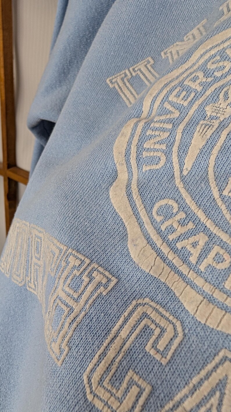 May include: Pale blue sweatshirt featuring a white graphic. The graphic includes the words "UNIVERSITY" and "CHAPEL HILL" in a circular design. The sweatshirt is made of a soft, textured fabric.