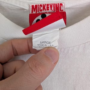 Puede incluir: Primer plano de una etiqueta de camiseta blanca con el texto "LARGE MADE IN U.S.A.". La etiqueta est&aacute; sujeta por una mano. La camiseta tiene una etiqueta de Mickey Inc.