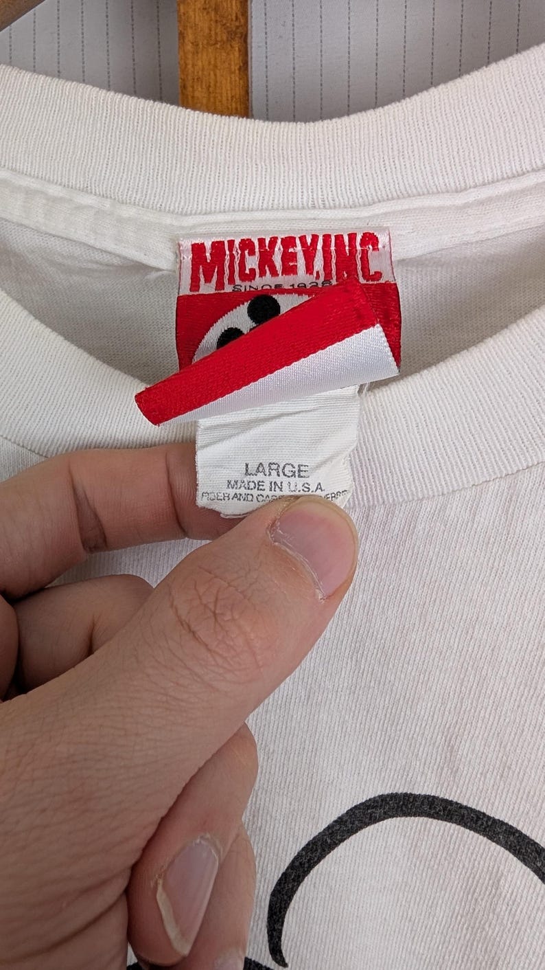Puede incluir: Primer plano de una etiqueta de camiseta blanca con el texto "LARGE MADE IN U.S.A.". La etiqueta est&aacute; sujeta por una mano. La camiseta tiene una etiqueta de Mickey Inc.
