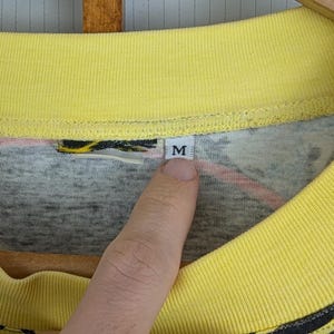 Pu&ograve; includere: Primo piano di una maglietta con colletto giallo con un design grigio e nero. La maglietta ha un'etichetta con la lettera "M". La maglietta &egrave; appesa a una gruccia di legno.