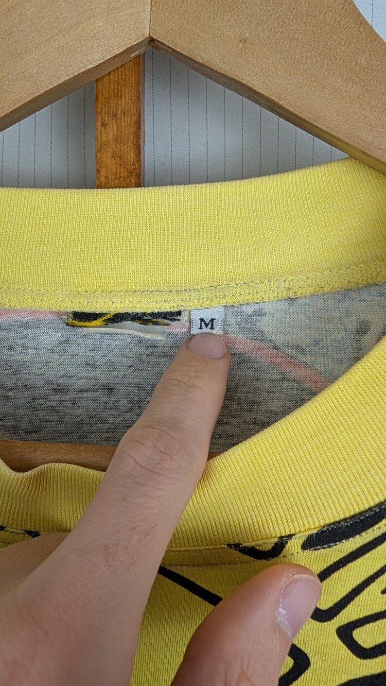 Pu&ograve; includere: Primo piano di una maglietta con colletto giallo con un design grigio e nero. La maglietta ha un'etichetta con la lettera "M". La maglietta &egrave; appesa a una gruccia di legno.