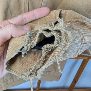 Peut inclure: Gros plan sur une manche de veste Carhartt beige, montrant des effilochages et des dommages importants au niveau du poignet. Le logo Carhartt est visible sur le haut de la veste. La doublure int&eacute;rieure de la manche est expos&eacute;e.