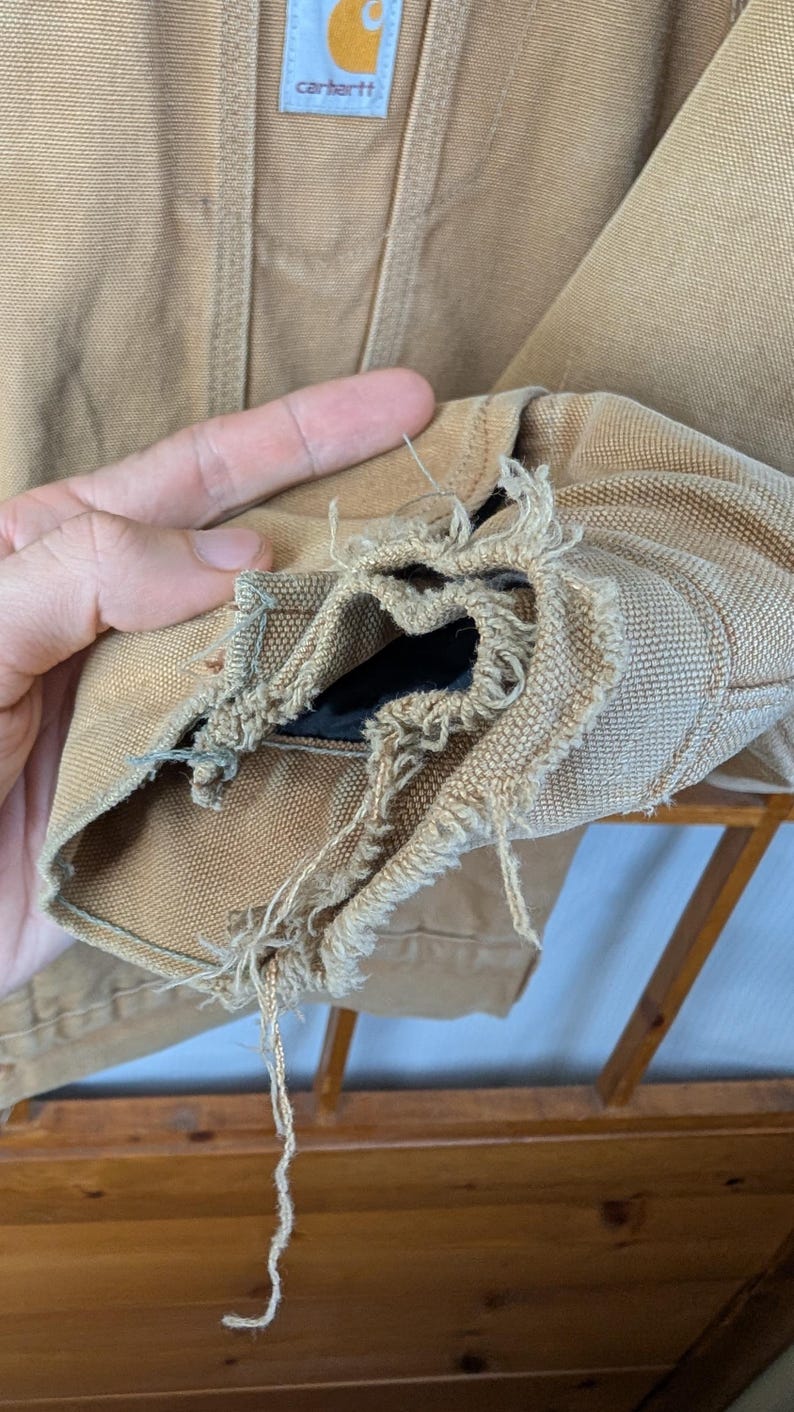 Peut inclure: Gros plan sur une manche de veste Carhartt beige, montrant des effilochages et des dommages importants au niveau du poignet. Le logo Carhartt est visible sur le haut de la veste. La doublure int&eacute;rieure de la manche est expos&eacute;e.
