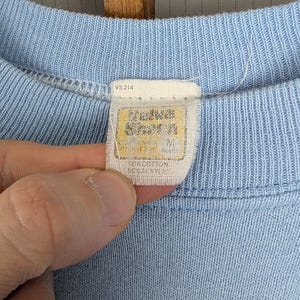 May include: Pale blue crew neck shirt with a tag that reads "Andrews Seven" and "Size M". The tag also indicates the shirt is made of 50% cotton and 50% acrylic. The shirt is a solid pale blue colour.