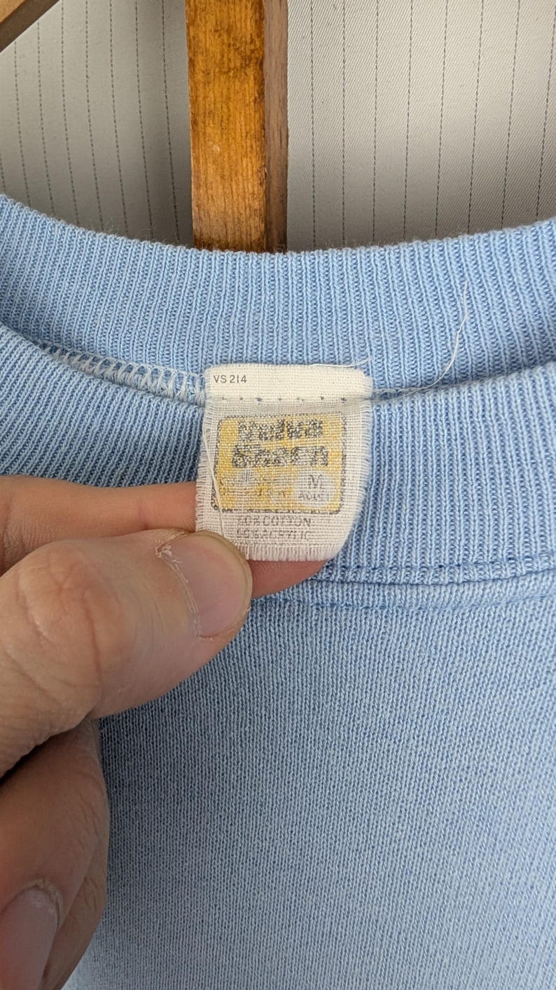 May include: Pale blue crew neck shirt with a tag that reads "Andrews Seven" and "Size M". The tag also indicates the shirt is made of 50% cotton and 50% acrylic. The shirt is a solid pale blue colour.