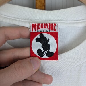 Puede incluir: Camiseta blanca con un dise&ntilde;o de Mickey Mouse. La etiqueta dice "MICKEY INC SINCE 1928" con una silueta negra de Mickey Mouse dentro de un c&iacute;rculo blanco sobre un fondo rojo. La camiseta est&aacute; colgada en una percha de madera.