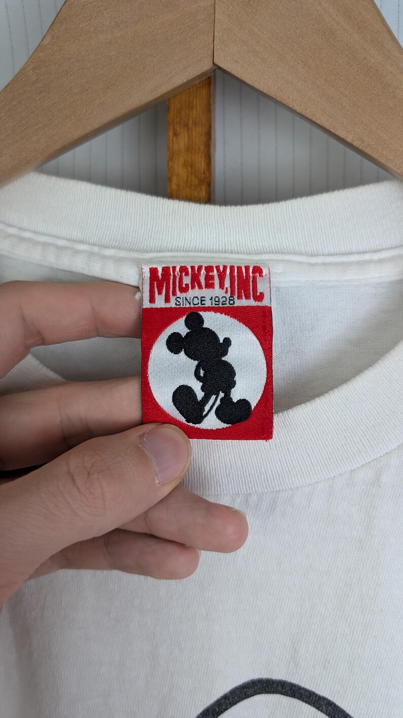 Puede incluir: Camiseta blanca con un dise&ntilde;o de Mickey Mouse. La etiqueta dice "MICKEY INC SINCE 1928" con una silueta negra de Mickey Mouse dentro de un c&iacute;rculo blanco sobre un fondo rojo. La camiseta est&aacute; colgada en una percha de madera.