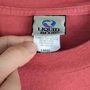 Puede incluir: Primer plano de una camiseta roja con una etiqueta Liquid Blue. La etiqueta incluye instrucciones de cuidado y la talla "LARGE". La tela de la camiseta es visible y la etiqueta est&aacute; sujeta por una mano.