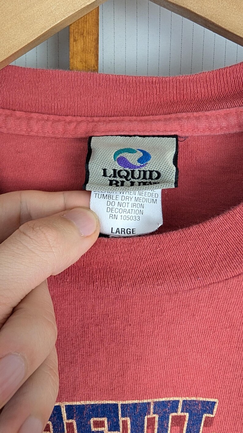 Puede incluir: Primer plano de una camiseta roja con una etiqueta Liquid Blue. La etiqueta incluye instrucciones de cuidado y la talla "LARGE". La tela de la camiseta es visible y la etiqueta est&aacute; sujeta por una mano.