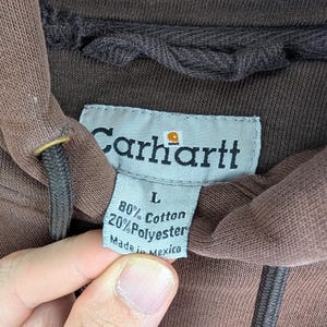 May include: Close-up of a brown Carhartt hoodie label. The label reads "Carhartt" with the brand's logo, and lists the fabric composition as 80% cotton and 20% polyester. The label also indicates the size is L and "Made in Mexico."