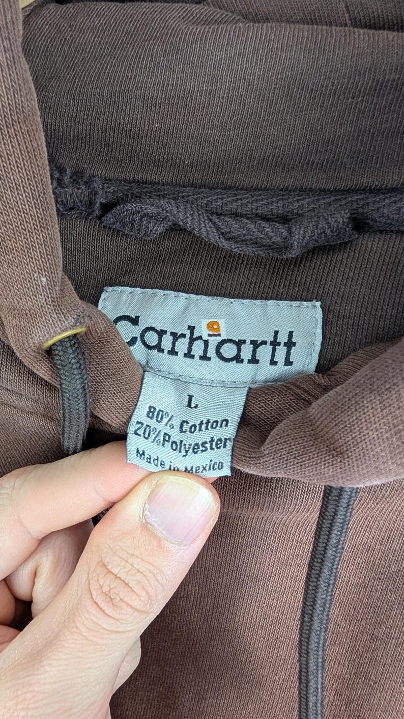 May include: Close-up of a brown Carhartt hoodie label. The label reads "Carhartt" with the brand's logo, and lists the fabric composition as 80% cotton and 20% polyester. The label also indicates the size is L and "Made in Mexico."