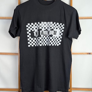 May include: Black t-shirt with a white checkered design and the text "NEW WAVE WEDNESDAY THE DEVILHOUSE". The shirt is hanging on a wooden hanger against a wooden background.