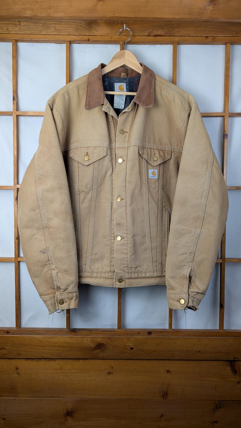 Peut inclure: Veste de travail beige avec un col en velours c&ocirc;tel&eacute; marron, une fermeture boutonn&eacute;e et deux poches poitrine. La veste a des boutons dor&eacute;s et un &eacute;cusson logo Carhartt. La veste est suspendue &agrave; un cintre en bois.