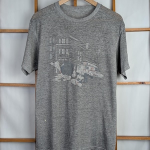 May include: A grey short-sleeved t-shirt featuring a graphic print of a building and bridge scene. The design is in shades of grey and white, with red accents. The t-shirt is hanging on a wooden hanger.