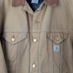 Peut inclure: Veste Carhartt beige avec un col en velours c&ocirc;tel&eacute; marron, une fermeture boutonn&eacute;e et deux poches poitrine. La veste a une doublure matelass&eacute;e et le logo Carhartt est visible sur une petite &eacute;tiquette. La veste est suspendue &agrave; un cintre en bois.