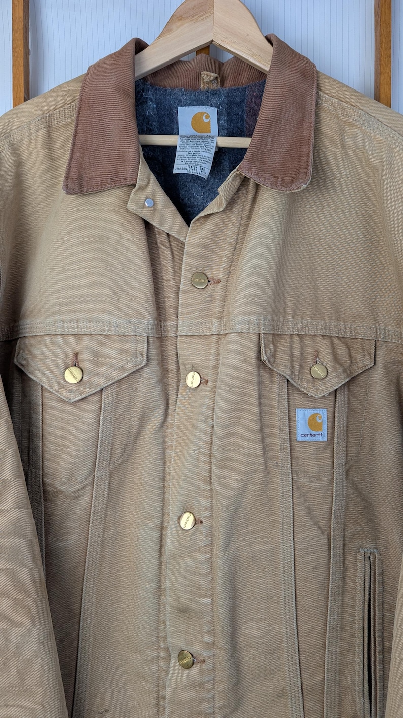 Peut inclure: Veste Carhartt beige avec un col en velours c&ocirc;tel&eacute; marron, une fermeture boutonn&eacute;e et deux poches poitrine. La veste a une doublure matelass&eacute;e et le logo Carhartt est visible sur une petite &eacute;tiquette. La veste est suspendue &agrave; un cintre en bois.