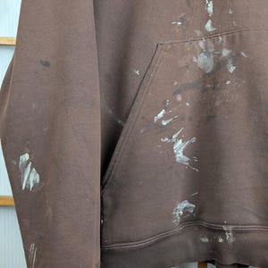 May include: A brown hoodie with a front pocket and ribbed hem and cuffs. The hoodie has white and grey paint splatters on the front and sleeves. The fabric appears to be a soft, brushed material.