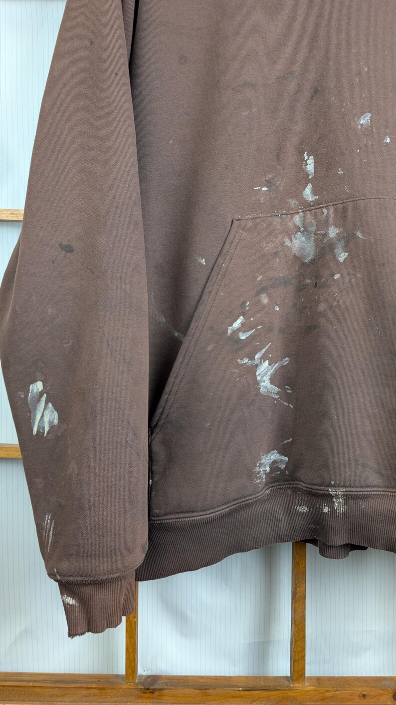 May include: A brown hoodie with a front pocket and ribbed hem and cuffs. The hoodie has white and grey paint splatters on the front and sleeves. The fabric appears to be a soft, brushed material.