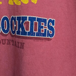 Puede incluir: Camiseta de color rosa polvoriento con un gr&aacute;fico desgastado. El gr&aacute;fico presenta la palabra "ROCKIES" en letras de bloque azules y blancas, con un dise&ntilde;o de cordillera amarilla encima. La palabra "MOUNTAIN" est&aacute; debajo.