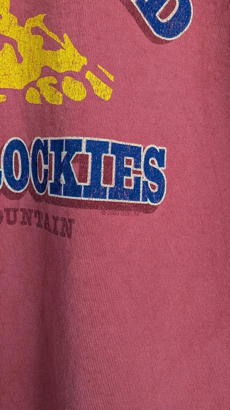 Puede incluir: Camiseta de color rosa polvoriento con un gr&aacute;fico desgastado. El gr&aacute;fico presenta la palabra "ROCKIES" en letras de bloque azules y blancas, con un dise&ntilde;o de cordillera amarilla encima. La palabra "MOUNTAIN" est&aacute; debajo.