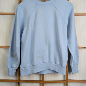 May include: A pale blue sweatshirt with long sleeves and a crew neck, hanging on a wooden hanger. The background is a wooden window frame. The sweatshirt has raglan sleeves.