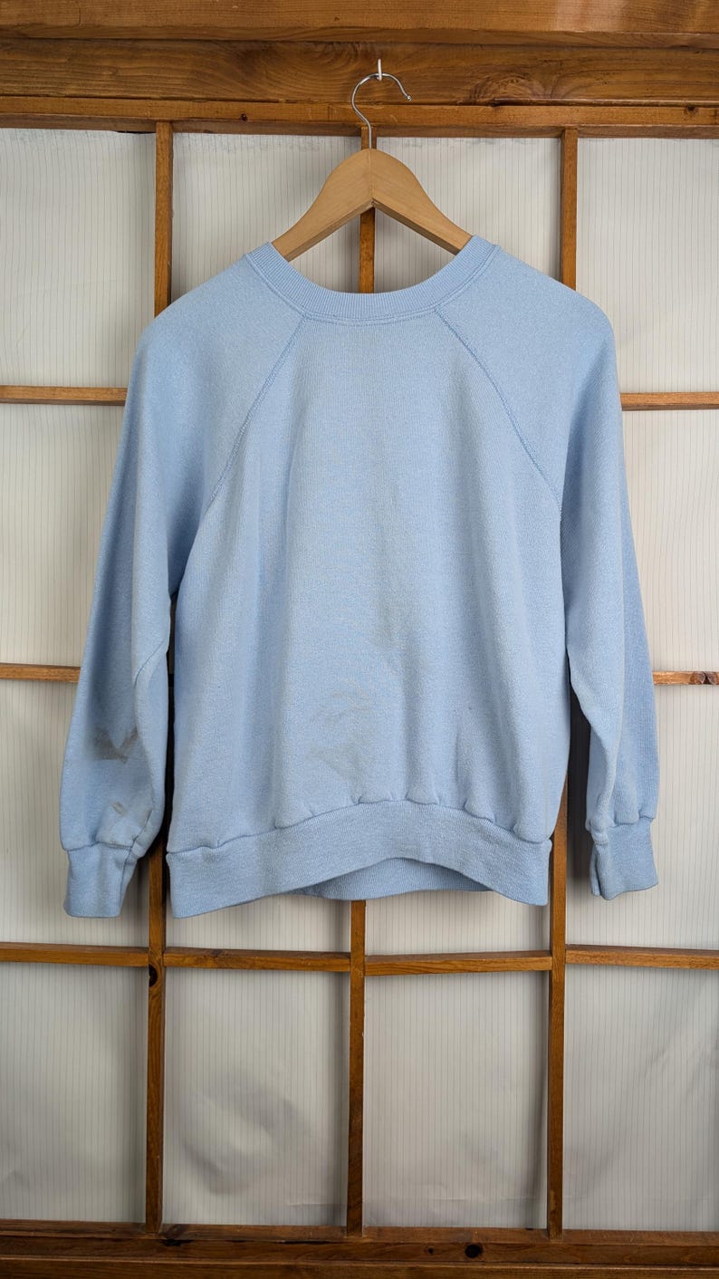 May include: A pale blue sweatshirt with long sleeves and a crew neck, hanging on a wooden hanger. The background is a wooden window frame. The sweatshirt has raglan sleeves.