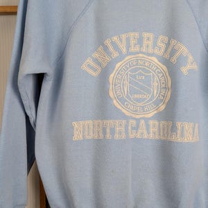 May include: Pale blue sweatshirt featuring the text "UNIVERSITY OF NORTH CAROLINA CHAPEL HILL" in white. The garment has a crew neck and long sleeves. The university seal is also printed in white.