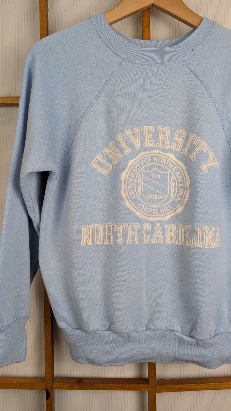May include: Pale blue sweatshirt featuring the text "UNIVERSITY OF NORTH CAROLINA CHAPEL HILL" in white. The garment has a crew neck and long sleeves. The university seal is also printed in white.