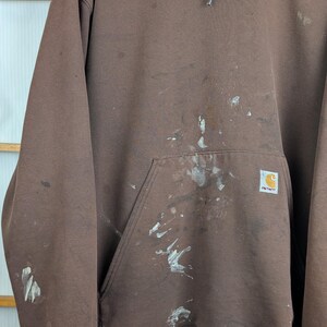 May include: A brown Carhartt hoodie with a drawstring hood and a kangaroo pocket. The hoodie has white paint splatters on the front and a Carhartt logo patch. The cuffs and hem are ribbed.
