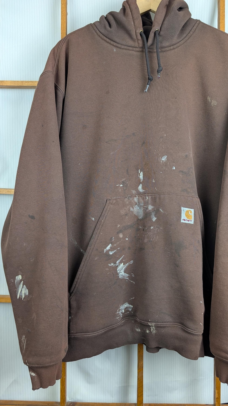 May include: A brown Carhartt hoodie with a drawstring hood and a kangaroo pocket. The hoodie has white paint splatters on the front and a Carhartt logo patch. The cuffs and hem are ribbed.