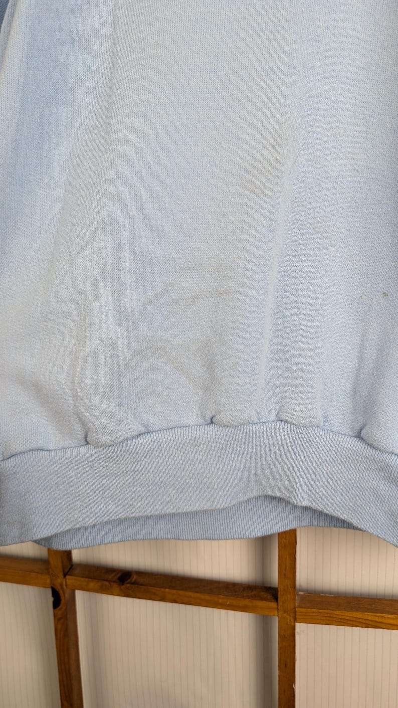 May include: Pale blue sweatshirt with a ribbed hem and cuffs. The fabric has a soft texture and some visible marks. The bottom hem is a darker shade of blue. A casual garment.