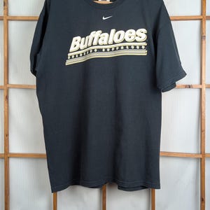Może przedstawiać: Czarny t-shirt z napisem "Buffaloes" w białych i złotych literach. Logo Nike jest widoczne nad tekstem. Koszulka wisi na drewnianym wieszaku.