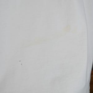 Peut inclure: Gros plan sur un tissu blanc l&eacute;g&egrave;rement textur&eacute;. Le tissu pr&eacute;sente quelques petites taches sombres et une tache plus grande de couleur claire. Le tissu est pli&eacute;, montrant le bord du mat&eacute;riau.