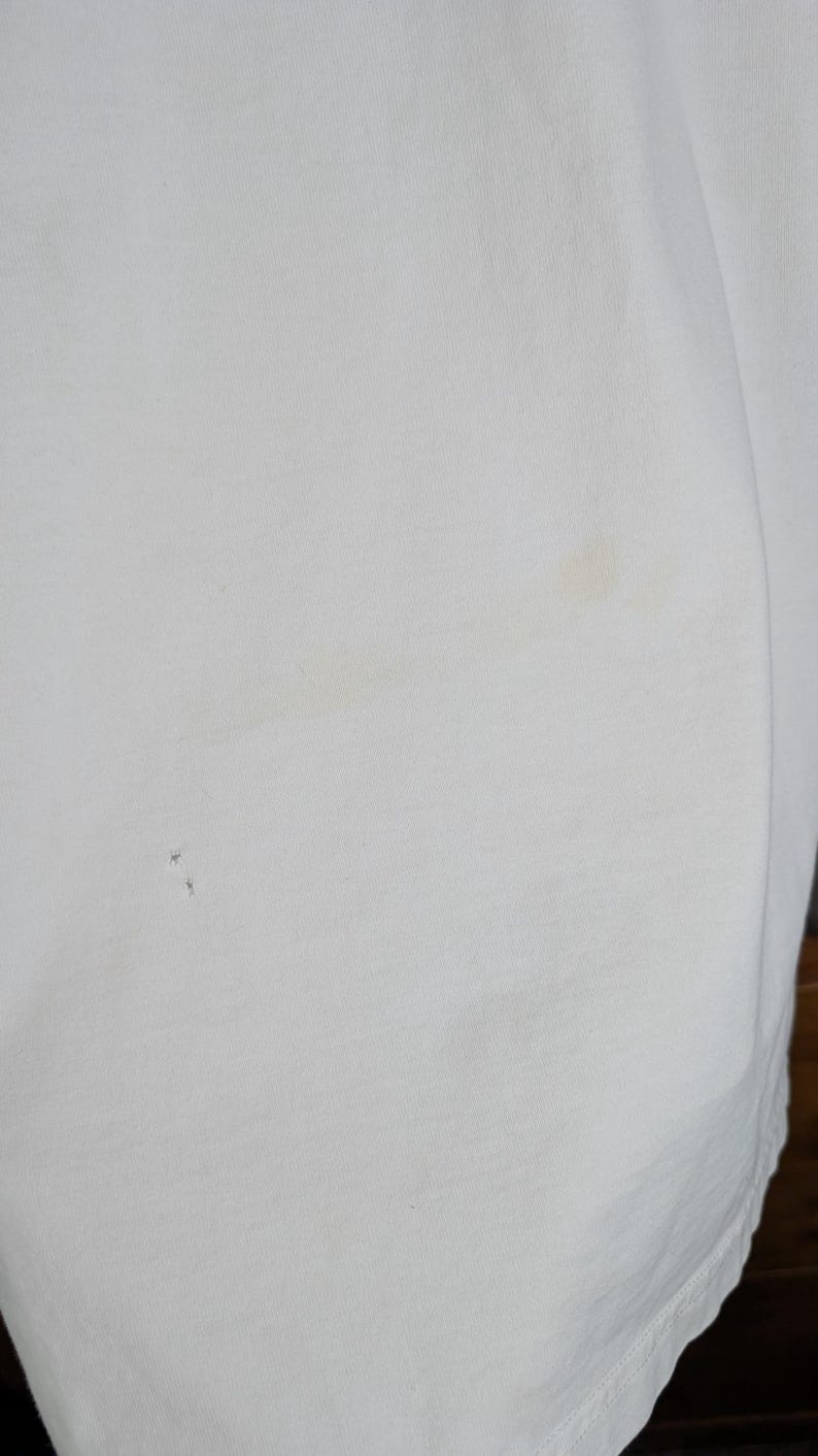 Peut inclure: Gros plan sur un tissu blanc l&eacute;g&egrave;rement textur&eacute;. Le tissu pr&eacute;sente quelques petites taches sombres et une tache plus grande de couleur claire. Le tissu est pli&eacute;, montrant le bord du mat&eacute;riau.