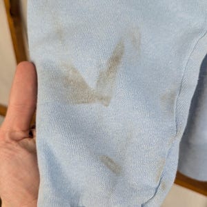 May include: Close-up of a pale blue sweatshirt sleeve with a ribbed cuff. The sleeve has several dark brown stains on the fabric. The background is out of focus.