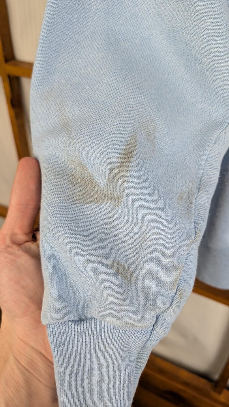 May include: Close-up of a pale blue sweatshirt sleeve with a ribbed cuff. The sleeve has several dark brown stains on the fabric. The background is out of focus.