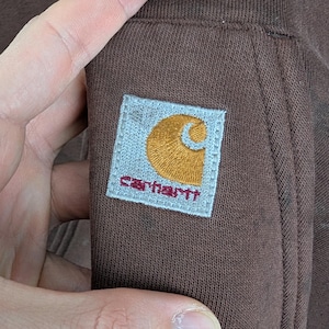 May include: Close-up of a brown Carhartt sweatshirt. The Carhartt logo patch is visible, featuring a gold 'C' and the brand name in red on a light blue square. The fabric is a solid brown colour.