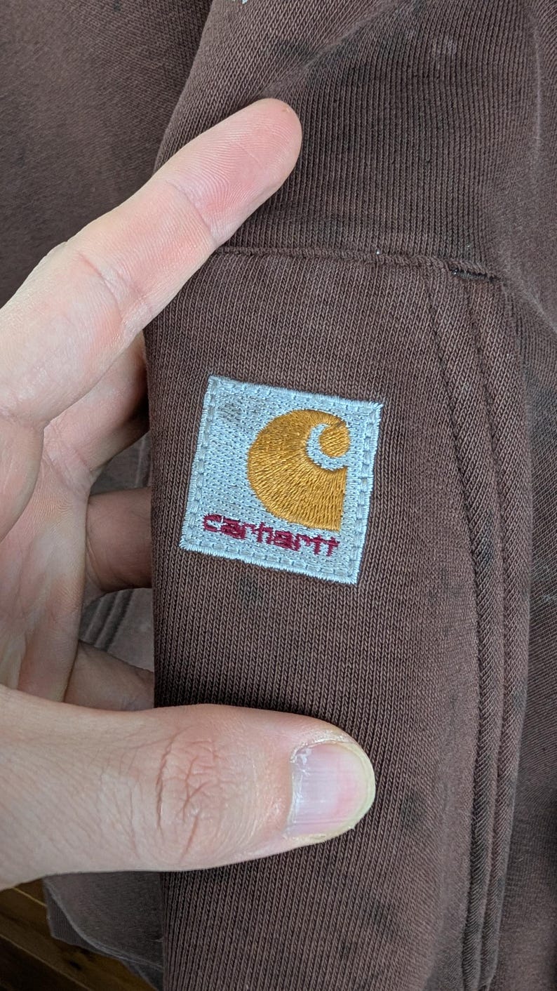May include: Close-up of a brown Carhartt sweatshirt. The Carhartt logo patch is visible, featuring a gold 'C' and the brand name in red on a light blue square. The fabric is a solid brown colour.