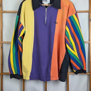 Puede incluir: Una camisa de manga larga con bloques de color con cuello y puños negros. La camisa presenta paneles amarillos, morados, naranjas y negros, con una manga de rayas arcoíris. La palabra "YES" está impresa en el cuello y el pecho.