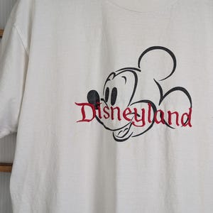 Puede incluir: Camiseta blanca con un contorno negro de Mickey Mouse y la palabra "Disneyland" en escritura roja. La camiseta tiene cuello redondo y una etiqueta "Mickey Inc.". Un tema cl&aacute;sico de Disney.
