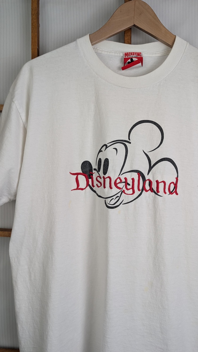 Puede incluir: Camiseta blanca con un contorno negro de Mickey Mouse y la palabra "Disneyland" en escritura roja. La camiseta tiene cuello redondo y una etiqueta "Mickey Inc.". Un tema cl&aacute;sico de Disney.