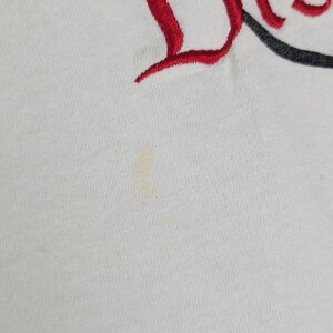 Puede incluir: Camiseta blanca con la palabra "Dis" bordada en rojo y un dise&ntilde;o de oreja de Mickey Mouse negro. La camiseta tiene una peque&ntilde;a mancha en la parte delantera.