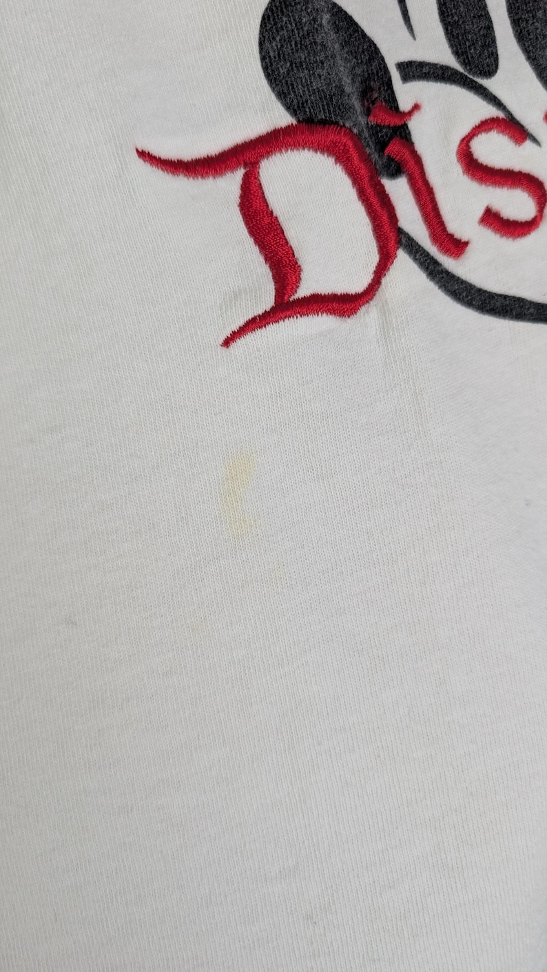 Puede incluir: Camiseta blanca con la palabra "Dis" bordada en rojo y un dise&ntilde;o de oreja de Mickey Mouse negro. La camiseta tiene una peque&ntilde;a mancha en la parte delantera.