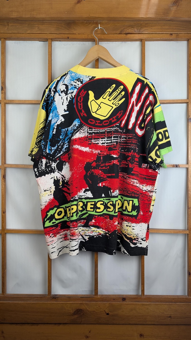 Pu&ograve; includere: Una maglietta vintage e colorata con un design grafico audace. La maglietta presenta un colletto e maniche gialle, con un vivace mix di arte astratta rossa, nera, bianca e blu. Il testo "BODY GLOVE" e "OPPRESSSN" &egrave; visibile.