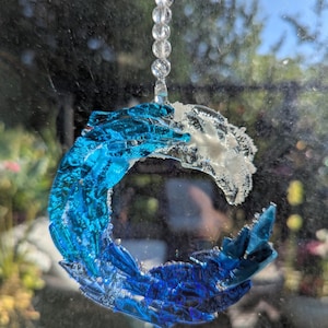 Puede incluir: Un atrapasoles decorativo con forma de ola, hecho de vidrio azul y blanco. La ola está suspendida por una cadena de cuentas transparentes. El diseño está pensado para captar la luz y añadir un toque costero al hogar.
